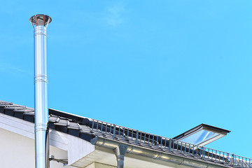 Stainless steel chimney and parts of a roof with an open roof window in front of a bright blue sky.