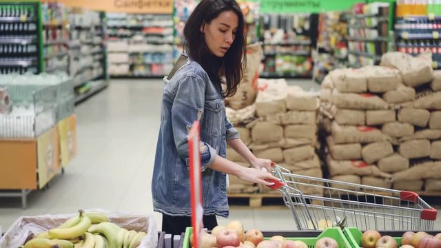 Attractive girl happy customer is choosing fruit in supermarket buying bananas, apples and oranges and putting them in shopping cart. Healthy lifestyle and shops concept.