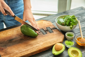 Young woman cutting avocado for nourishing mask at table