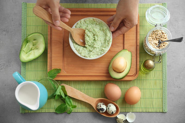 Young woman making nourishing mask with avocado at table
