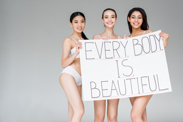 smiling multicultural women holding banner with lettering every body is beautiful isolated on gray background