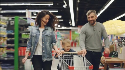 Slow motion of happy family father, mother and child running through supermarket with shopping cart, smiling and laughing. Having fun in shop and people concept. - Powered by Adobe