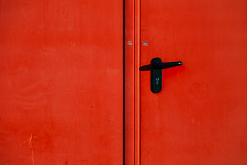 The modern door with a black handle and a lock of red color is closed. Log into the container or mobile home. Red wall. Cobbles or tiles. Three door hinges. Sunset shadow. Close up view