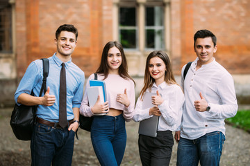 Cheerful four friends are standing near the university. They are giving thumbs up with happiness.