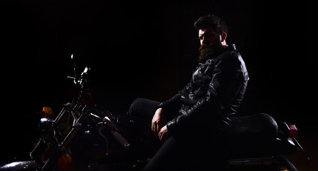 Fototapeta na wymiar Man with beard, biker in leather jacket sitting on motor bike in darkness, black background. Macho, brutal biker in leather jacket riding motorcycle at night time, copy space. Night racer concept.
