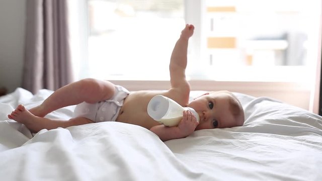 Pretty baby girl drinks water from bottle lying on bed. Child weared diaper