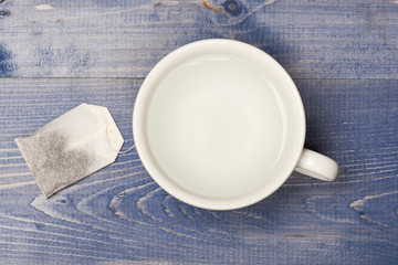 Cup or white mug with transparent hot water and bag of tea. Tea time concept. Mug filled with boiling water and teabag on blue wooden background. Process of tea brewing in ceramic mug, top view