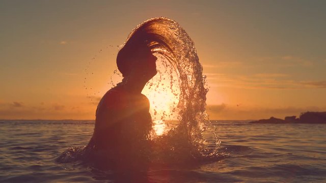 SLOW MOTION, CLOSE UP, SILHOUETTE, LENS FLARE: Young woman whips her hair back and splashes water over the sunset. Unrecognizable girl in bikini plays in the refreshing ocean water in the evening.