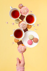 Obraz na płótnie Canvas Cups of tea, pink, delicious cupcakes, colorful macaroons and chocolate pieces are balancing on the index finger. Modern conceptual photography on a yellow background.