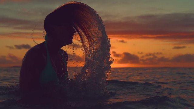 SLOW MOTION, CLOSE UP, SILHOUETTE: Young woman flips her long hair back and splashes glassy water on a breathtaking evening on exotic beach. Playful female traveler cools off in the refreshing sea.