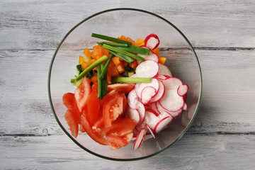Glass bowl with vegetables for a vegetarian salad. Radishes, tomatoes, celery, bell pepper, onion and cucumber. White wooden kitchen table. Close up. Top view.