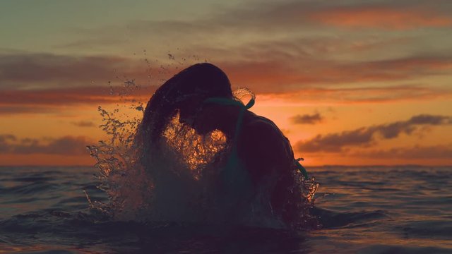 SLOW MOTION SILHOUETTE CLOSE UP: Unrecognizable girl whips her head out of the ocean and splashes glistening water over the orange lit sunset. Energetic young woman splashing sea water with her hair.
