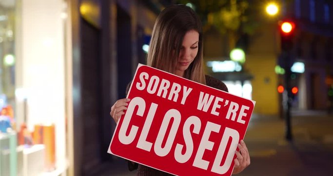 Woman holding Closed sign outside her small shop in the city at night, Caucasian Female millennial looking at camera with Sorry We Are Closed sign outside store on sidewalk, 4k
