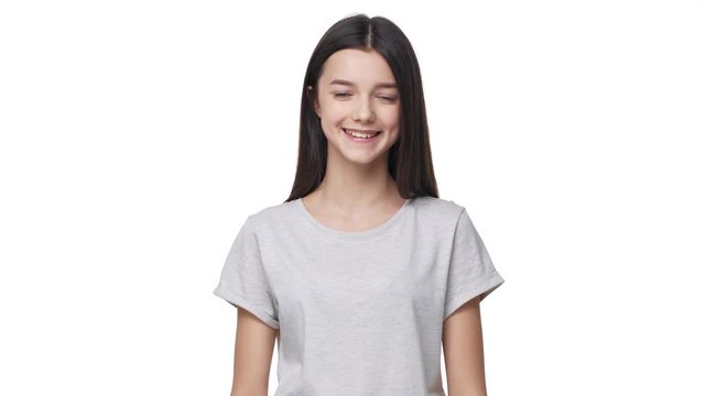 Portrait of beautiful teen girl in basic t-shirt smiling at camera and touching her long dark hair, isolated over white background in studio. Concept of emotions