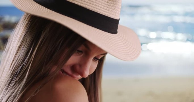 Close-up of woman wearing hat looking over her shoulder in sunny beach setting, Pretty female in her 20s looking at camera and smiling by the ocean outdoors, 4k