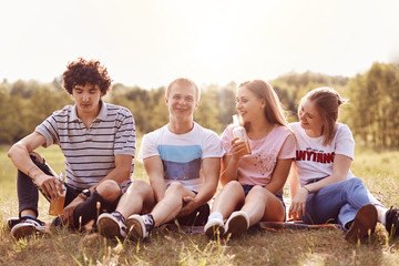 Fototapeta premium Four positive friends have joy together, have happy expressions, sit on green grass outdoor, enjoy spare time during summer vacation, pose outdoor at nature. People, friendship and recreation concept