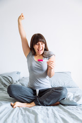 young happy woman sitting on bed looking on positive pregnancy test