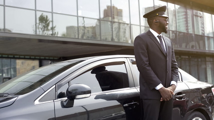 Cheerful Afro-American taxi service driver expecting passengers near luxury car