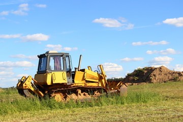 Yellow tractor (bulldozer with a plow) closeup on a green field on the background of the blade of the earth day in the summer on a background of blue sky with clouds