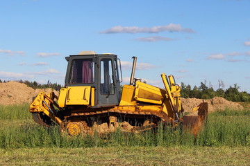 Yellow tractor (bulldozer with a plow) closeup on a green field on the background of the blade of the earth day in the summer on a background of blue sky with clouds