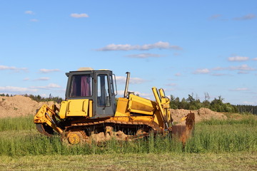 Yellow tractor (bulldozer with a plow) closeup on a green field on the background of the blade of the earth day in the summer on a background of blue sky with clouds