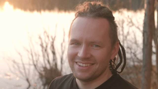 Young man with dreadlocks looking at the camera and smiling. Stands on the riverbank. Close-up face.