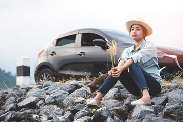 Young asian woman sitting with chihuahua dogs beside the road with black hatchback eco car. Stylish attractive girl traveler wear denim shirt traveling with pets in summer vacation. Lifestyle outdoor