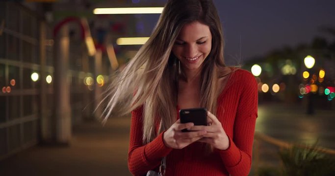 Millennial woman texting on smartphone looking happy outside in street in the evening, Caucasian girl in her 20s happily texting on phone standing on sidewalk at night, 4k