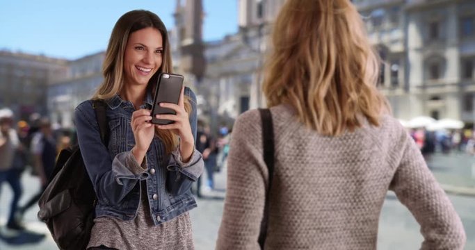Beautiful young brunette taking photo of pretty blonde friend in Rome, Italy, Cheerful white female takes picture of posing friend using her smartphone, 4k