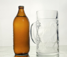 A bottle of fresh beer, still plugged and with drops of water on the glass surface, and a giant transparent mug. White background.
