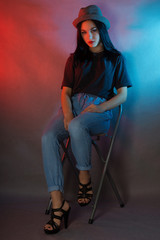 Young girl in blue jeans and black t-shirt sitting on a chair on a blue-pink background