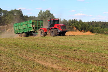 Obraz premium Agriculture, harvesting - new red tractor with green trailer carries silo on green field in summer afternoon on blue clear sky background with white clouds