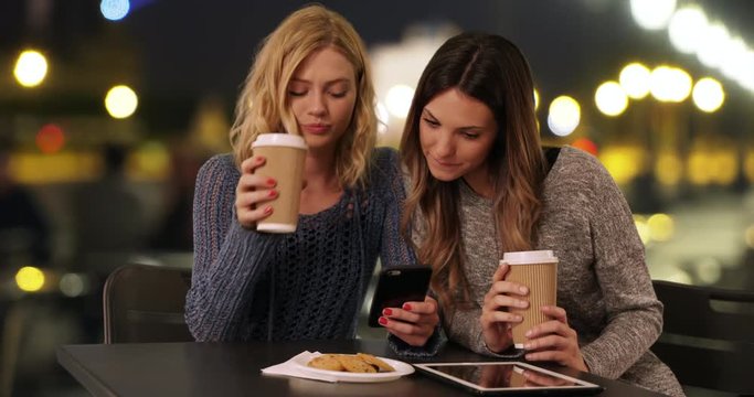 Couple of happy girls drinking together while looking at cell phone in Paris, France, Cute young caucasian women looking at smartphone outdoors French plaza, 4k