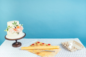Fototapeta na wymiar copyspace mockup working space baker confectioner pastry chef creamy white two-tiered wedding (birthday) cake with fresh flowers on table and cookies in studio on blue background