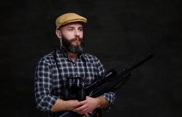 Fototapeta premium Portrait of a bearded hunter traveler in a fleece shirt and hat holds rifle with a sight and binoculars.