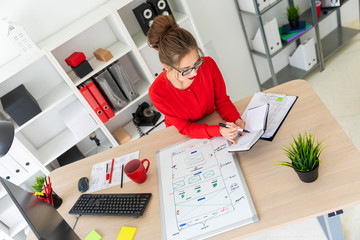 A young girl is sitting at the desk in the office, holding a black marker in her hand and working with a notepad. A magnetic board lies before the girl.