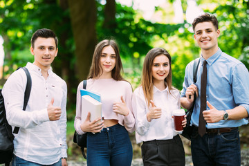 Four happy young people showing their thumbs up and smiling while standing close to each other outdoors