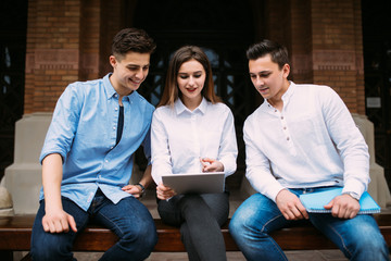 Group of young students sitting on bench in campus park and looking at tablet