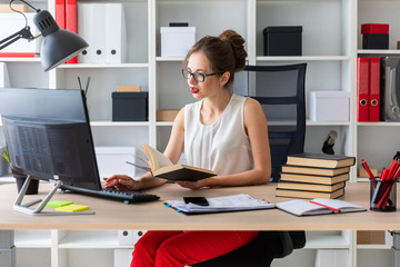 A young girl sits at a computer table and holds an open book in her hands.