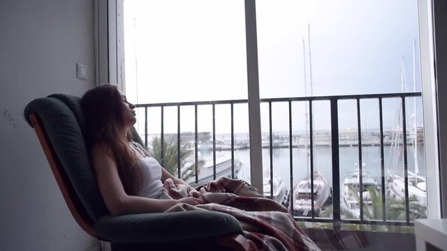 Young depressed woman swinging in a chair and looking out the window on sea. Raining outside. Lonely woman in despair.
