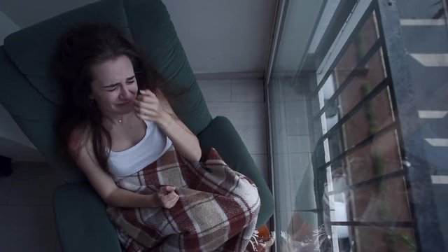 Top view of young depressed woman pulling her hair with hands and crying sitting in chair by the window. Raining outside. Lonely woman in despair with suicide thoughts.