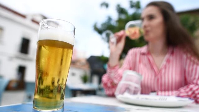 Close-up of bubbles and foam in a glass of light beer on the wooden table in the outdoors restaurant with young attractive woman drinking wine on background.