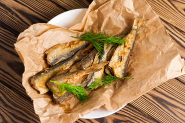 Heap of fried tasty fishes and twigs of fresh green dill on wrinkled paper on white ceramic plate on old wooden rustic brown table