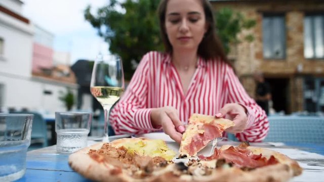 Slow motion of young woman taking a piece of pizza with hands and starting eating at outdoors restaurant. Freshly baked Italian pizza with prosciutto, mushrooms, artichokes, tuna and cheese.