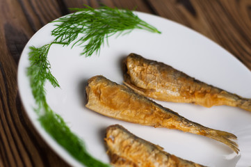 Row of three fried tasty fishes and twigs of fresh green dill on white ceramic dish on old wooden rustic brown table