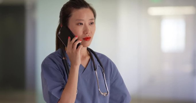 Working Millennial Nurse Dressed In Scrubs Talking On Cellphone Indoors Studio Clinic, Serious Young Asian Medical Practitioner Making Phone Call Inside Empty Hospital Hallway, 4k