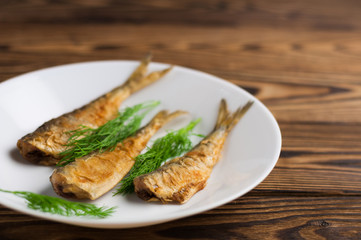 Row of three fried tasty fishes and twigs of fresh green dill on white ceramic dish on old wooden rustic brown table with copy space for your text