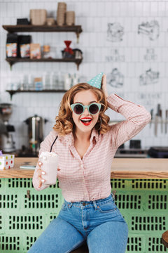 Cheerful lady in sunglasses and birthday cap sitting at the bar counter with milkshake in hand and happily spending time in cafe
