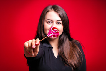 Cheerful fat brunette has fun posing with delicious lollipop. Plus size model, wear xxl bodysuit in the studio