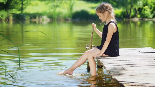Little girl in a black dress is sitting on a wooden pier and splashing with his feet in the water. Slow motion